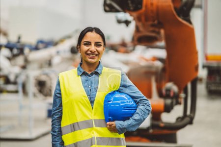 Smiling female engineer in safety gear holding a hard hat inside an industrial factory.