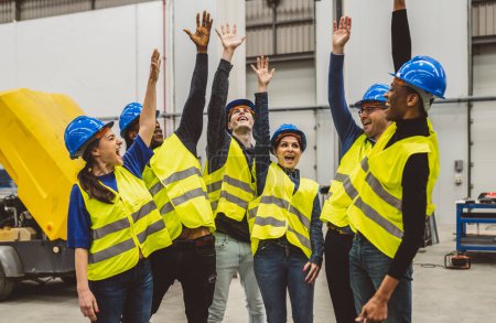 Grupo de ingenieros en equipo de seguridad celebrando con las manos levantadas en una fábrica industrial.