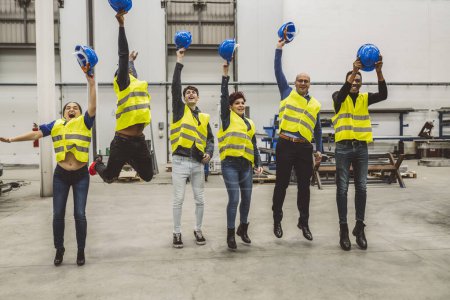 Gruppe von Ingenieuren in Sicherheitsausrüstung springt und wirft harte Hüte in die Luft, feiert in einer Industriefabrik.