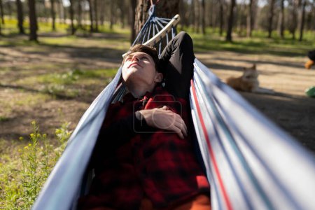 Jeune homme couché dans un hamac, profitant d'une journée ensoleillée dans la forêt avec un chien en arrière-plan.