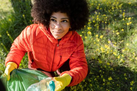 Jeune femme vêtue d'une veste rouge ramassant des déchets plastiques dans un parc, sensibilisant à l'environnement.