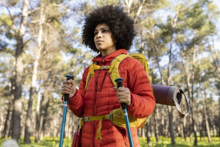 Young woman in a red jacket hiking with trekking poles and a backpack in a forest, looking determined.