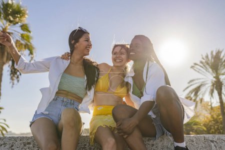 Trois jeunes femmes souriantes en bikinis et chemises blanches profitent d'une journée ensoleillée à la plage ensemble.