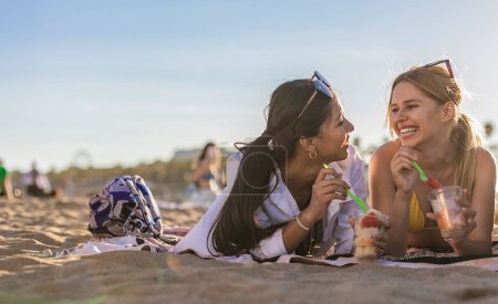 Zwei junge Frauen liegen am Strand, lachen und genießen Obstbecher an einem sonnigen Tag.