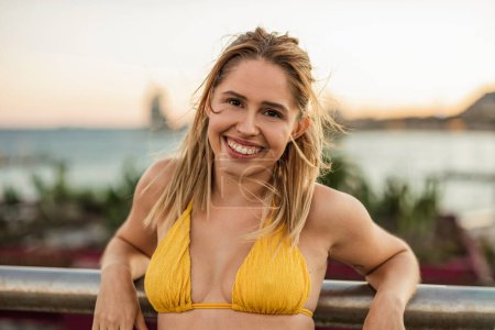 Téléchargez les photos : Jeune femme aux cheveux blonds, vêtue d'un bikini jaune, souriante et penchée sur une balustrade au bord de la plage au coucher du soleil. - en image libre de droit
