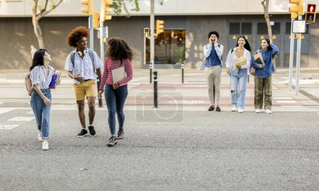 Eine Gruppe verschiedener College-Studenten überquert lachend die Straße und trägt an einem sonnigen Tag Bücher und Rucksäcke.