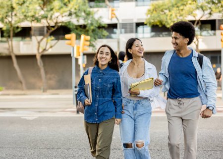 Three diverse college friends walking arm in arm, laughing and carrying books and backpacks on a sunny day.