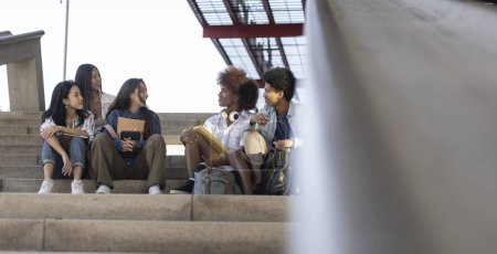 Group of diverse college students sitting on outdoor stairs, chatting and enjoying a sunny day together.