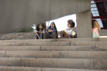 Diverse group of college students sitting on outdoor stairs, chatting and laughing, enjoying a sunny day.