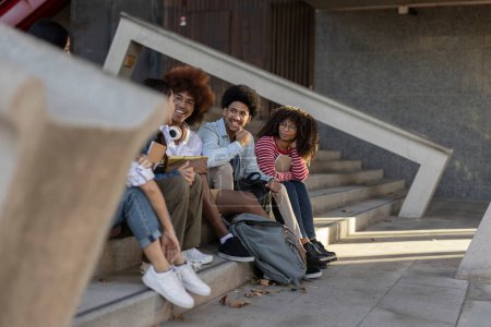 Group of diverse college students sitting on outdoor stairs, chatting and smiling, enjoying a sunny day on campus.