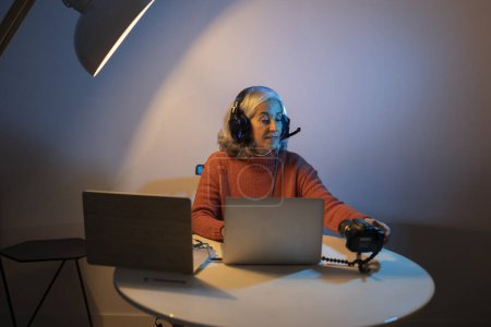 Mujer mayor con auriculares, trabajando en computadoras portátiles con una cámara en un escritorio, bajo iluminación de estudio.