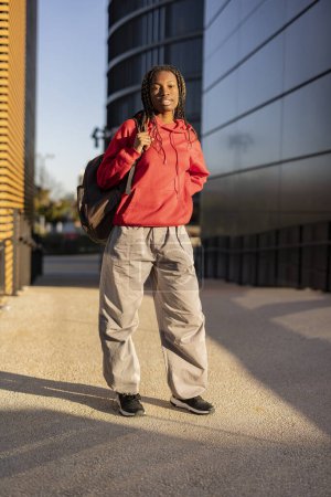 Selbstbewusste junge Frau mit rotem Kapuzenpullover und Rucksack, die in einem städtischen Umfeld mit modernen Gebäuden steht.