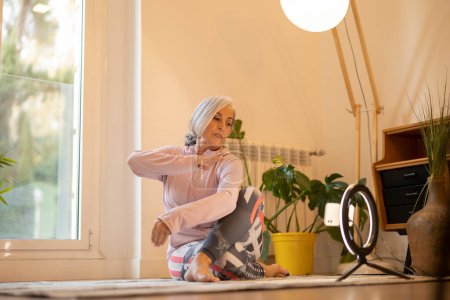 Femme âgée effectuant du yoga à la maison, avec un anneau lumineux et un smartphone pour l'enregistrement, assise sur un tapis.