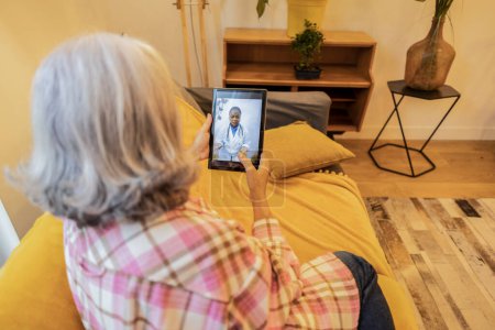 Elderly woman sitting on a couch, using a tablet for a telemedicine appointment with a doctor.