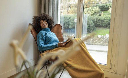 Young woman in blue sweater and yellow blanket relaxing and reading a book by a large window.