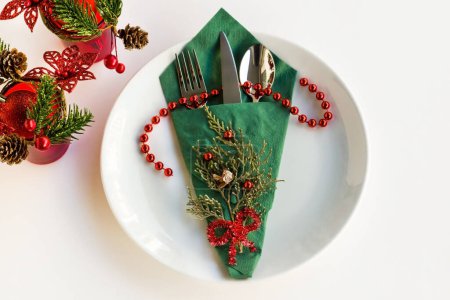 Téléchargez les photos : Serviette de Noël de couleur verte avec couverts fixés sur une assiette blanche à la surface blanche avec ornements de Noël - en image libre de droit