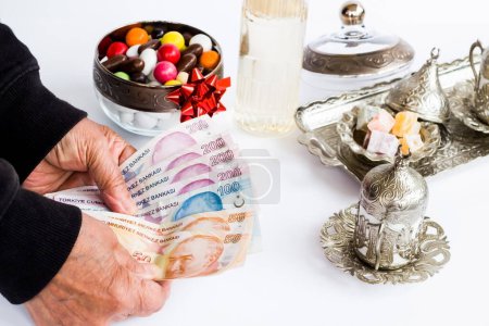Téléchargez les photos : Billets turcs en main de la personne âgée sur blanc avec de l'eau de Cologne, des bonbons colorés et un coffret de café turc. - en image libre de droit