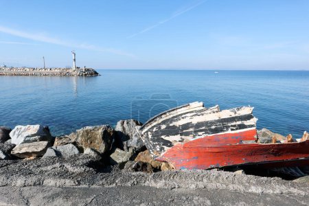 By the calm blue sea an old broken boat is lying on its side with a white lighthouse ahead