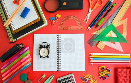 Téléchargez les photos : Fournitures scolaires colorées conçues sur une surface rouge, avec carnet à carreaux vierges et mini réveil vintage, Image conceptuelle de l'éducation - en image libre de droit