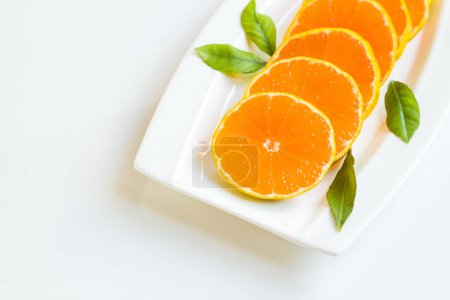 Téléchargez les photos : Mandarine fraîche, ronde, coupée en tranches avec des feuilles vertes fraîches, conçue sur une assiette blanche - en image libre de droit