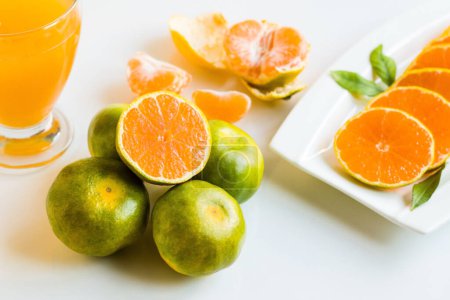 Téléchargez les photos : Les mandarines vertes et jaunes, dont l'une a été coupée en deux, ont été conçues sur un fond blanc avec un verre de jus de mandarine - en image libre de droit