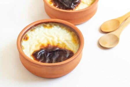 Téléchargez les photos : Pudding de riz cuit au four de style turc traditionnel dans un bol à casserole, sur surface blanche avec espace de copie et cuillère à dessert en bois - en image libre de droit