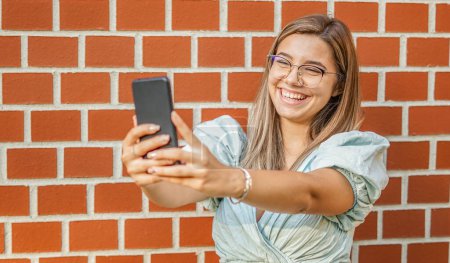 Foto de Young girl taking a selfie - Smiling at the camera - Happy student standing by the wall and using her smartphone - Imagen libre de derechos
