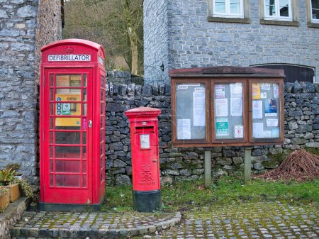Téléchargez les photos : Un défibrillateur d'urgence dans une vieille cabine téléphonique rouge dans le village de Taddington dans le Derbyshire, Royaume-Uni. Une boîte aux lettres rouge et un panneau d'affichage paroissial apparaissent à droite. - en image libre de droit