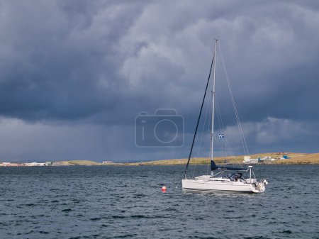 Foto de Un solo velero blanco atado a una boya en agua picada en Bressay Sound frente a Lerwick, Shetland, Reino Unido. El barco ondea la bandera de Shetland con Bressay en el fondo. - Imagen libre de derechos
