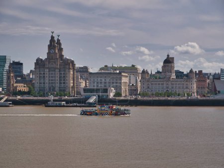 Photo for The River Mersey Ferry Snowdrop passes the Three Graces on the historic Liverpool waterfront. The art design is by Sir Peter Blake. - Royalty Free Image