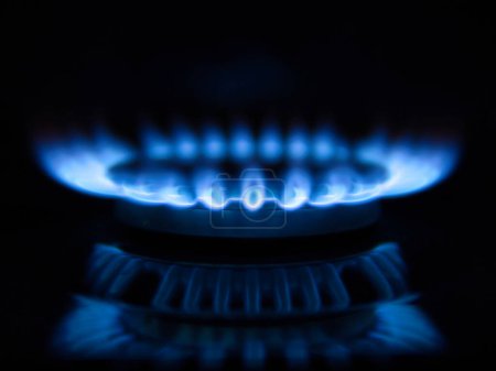 Les flammes bleues d'un brûleur à gaz sur une plaque de cuisson domestique, sur fond noir. Les flammes se reflètent dans l'émail noir de la plaque de cuisson.