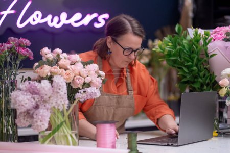 Téléchargez les photos : Fleuriste femme tapant sur ordinateur portable au comptoir de magasin de fleurs, vérifier les commandes de fleurs, petite entreprise - en image libre de droit