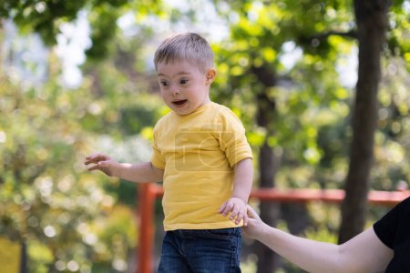 Loving mother holding hand of little son in yellow t-shirt to help climbing structure happy child with Down syndrome enjoys fun at children playground on sunny summer day