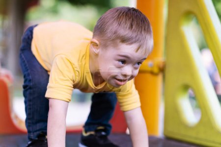 Foto de Niño aventurero con síndrome de Down preparándose para montar diapositivas en el patio lindo niño soleado en camiseta amarilla de pie en pose listo para divertirse en el día de verano - Imagen libre de derechos
