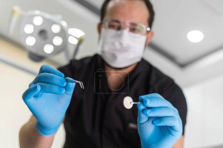 Male dentist in black uniform and gloves reaching to patient with mirror and tongs examining oral cavity at appointment in stomatology clinic triggering scene