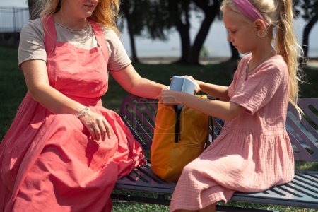 Téléchargez les photos : Écolière en robe rose met la boîte à lunch dans le sac à dos assis avec la jeune femme sur le banc du parc préoccupations de la mère au sujet du bien-être de la fille donnant de la nourriture pour le déjeuner à l'école - en image libre de droit