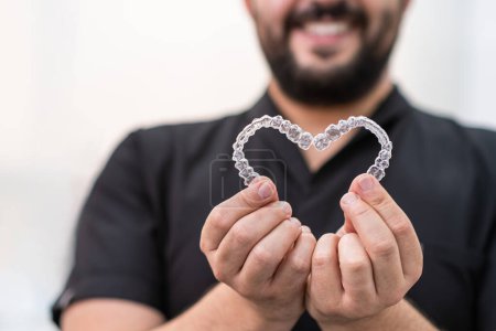 Smiling doctor holding heart made of clear aligners for professional teeth correction in stomatology clinic orthodontist showing love and care of patients closeup