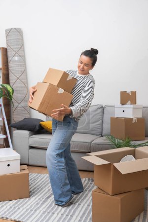Foto de Bienvenido a casa: Una mujer feliz, mudándose a su nuevo hogar, entra en la sala de estar con una pila de cajas móviles de cartón, dando la bienvenida al sentido de pertenencia - Imagen libre de derechos