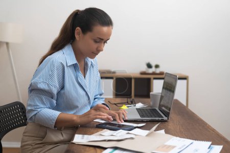 Serious lady calculates utility bills with calculator sitting at desk with laptop woman manages budget keeping track of bank calculations accuracy at home