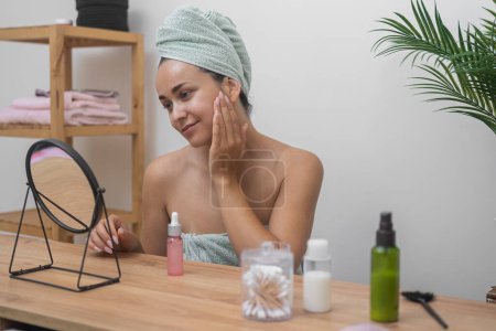 Foto de Serenidad en casa: Una mujer, con la cabeza envuelta en toallas, aplica suero de ácido hialurónico en la mesa espejada, elevando su rutina de cuidado de la piel. - Imagen libre de derechos