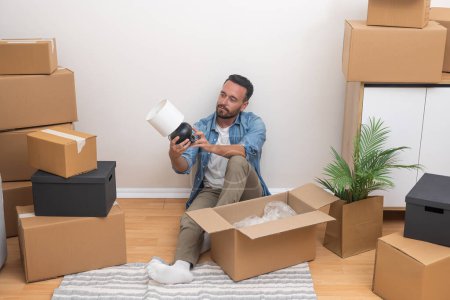 En medio de la reubicación, un joven desempacando una lámpara de entre cajas desempaquetadas en su nuevo apartamento, decidido a volver a casa