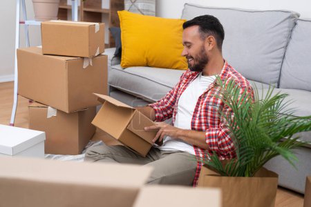 Ein junger Mann packt an seinem bewegenden Tag Kisten aus und findet einen Moment der Ruhe auf dem Sofa in seinem neuen Zuhause, das die Essenz des Umzugs symbolisiert