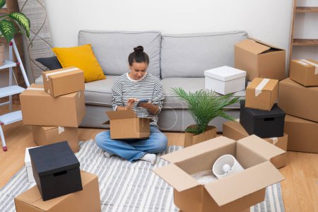 Moving Day Moments: Young woman unboxes on moving day, using her phone in new home, embodying the spirit of change