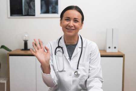 Foto de Consulta en línea Bienvenida: Con una sonrisa de bienvenida, una doctora saludando con la mano se involucra con la cámara, haciendo que los pacientes se sientan cómodos durante su cita en línea. - Imagen libre de derechos