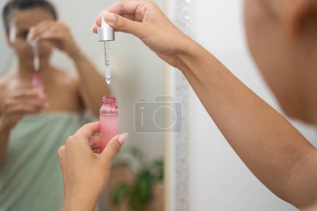 Foto de Mujer sosteniendo botella de suero cosmético con gotero cerca del espejo en el baño señora eligiendo ácido hialurónico para el tratamiento de la piel antienvejecimiento en el primer plano del hogar - Imagen libre de derechos