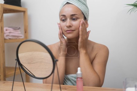 Foto de Espejo de alegría: Una señora, vestida de toalla, disfruta de un día de spa en el cuidado de la piel, admirando su suave piel en el espejo de la mesa. - Imagen libre de derechos