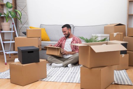 Young male with beard sits in new house unpacking things brought by courier in large cardboard boxes finding decorative items from old apartment with pleasant memories