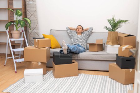 Woman sits on soft sofa waiting for help of husband in collecting boxes with furniture standing near wall with flower in pot with various shelves for books