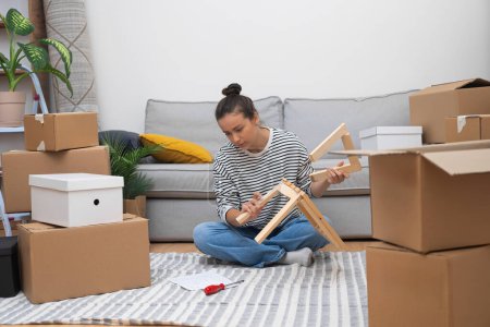 Rental Comfort: Woman tenant sits on the floor, assembling self-assembly shelf amidst moving carton boxes and furniture, making her new place feel like home