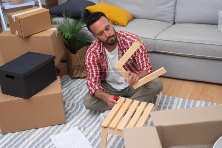 Building a New Life: A young man assembles a DIY shelf amidst moving carton boxes, adding warmth to his home after relocating